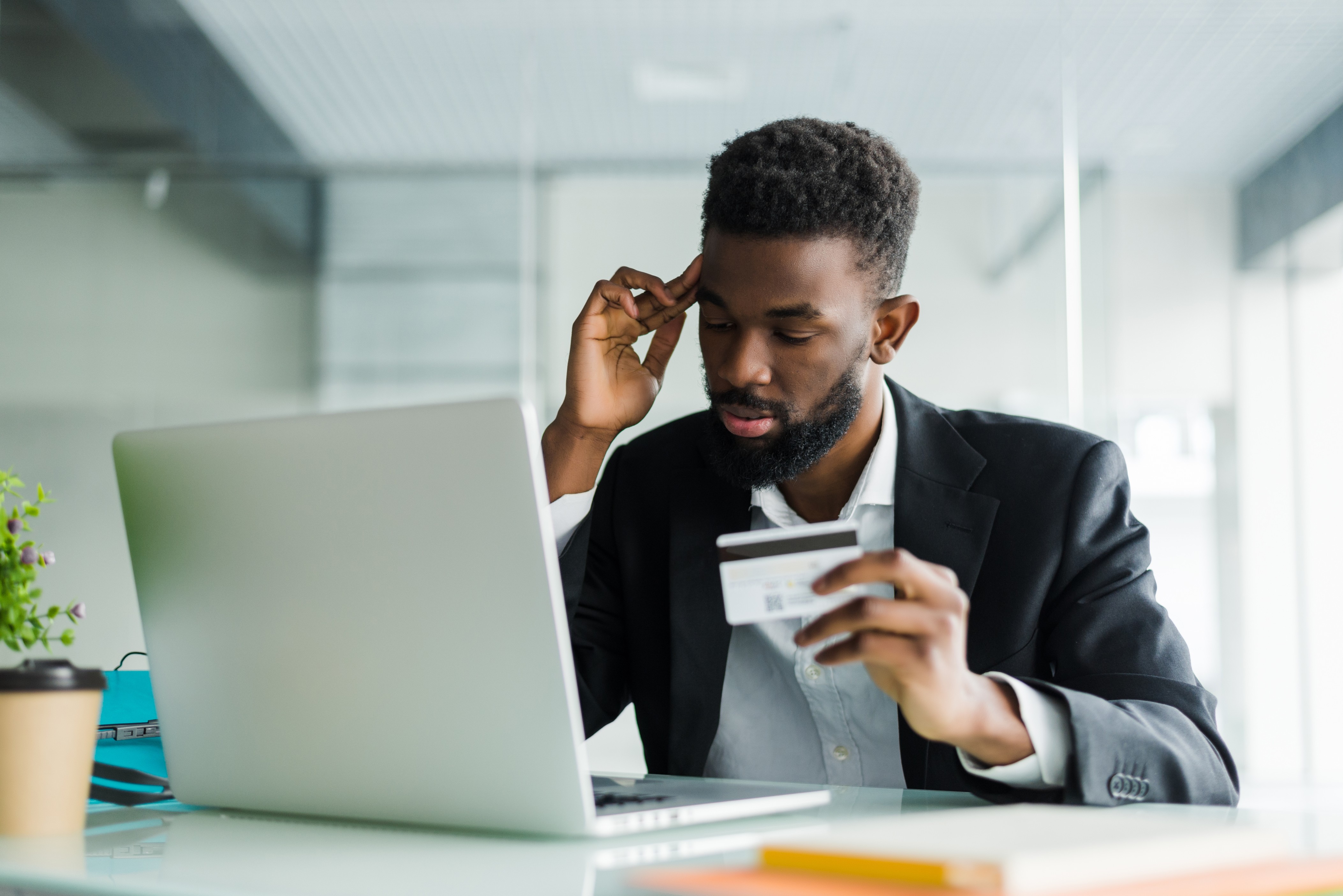 A young African man sitting at a desk, holding a credit card and looking at his laptop with a worried expression
