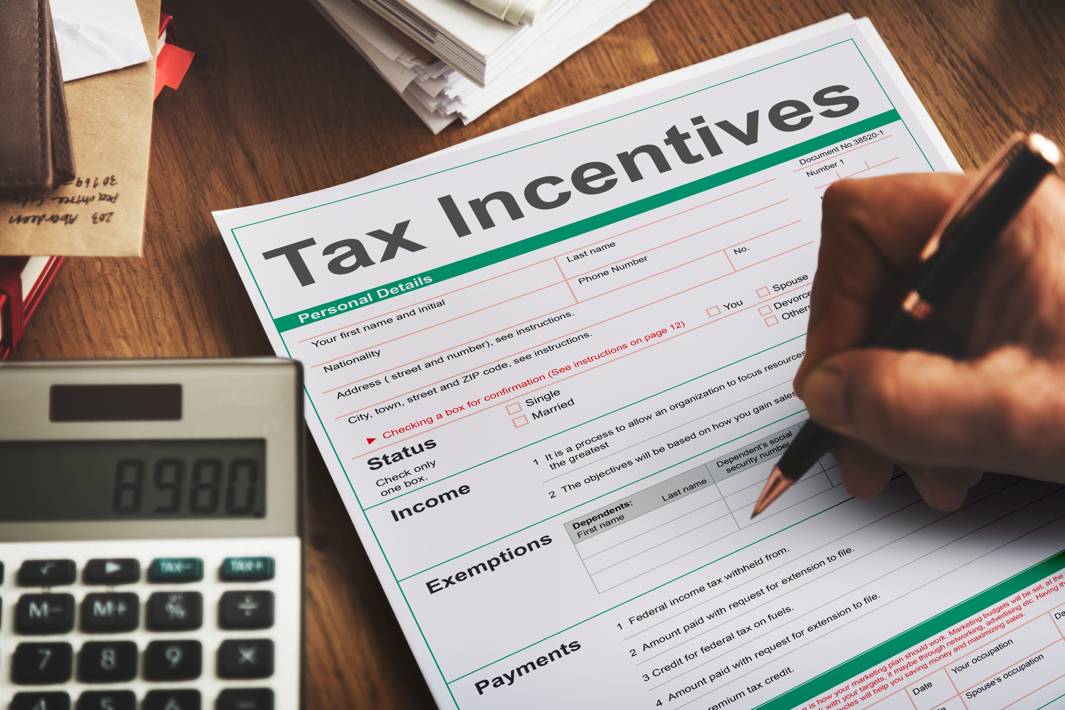 A person filling out a tax incentives form beside a calculator on a wooden desk